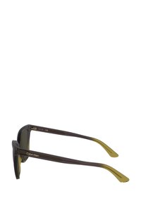 Ochelari de soare Calvin Klein cu lentile întunecate, un cadru transparent de culoare maro și accente galbene pe tâmple. Prezintă un design elegant și unghiular.