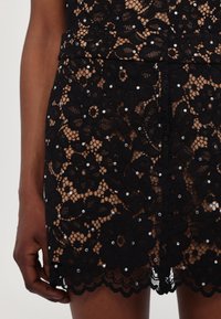 Jupe en dentelle noire avec motifs floraux et petites perles blanches, portée par une personne à la peau foncée, montrant la main et le haut de la cuisse.