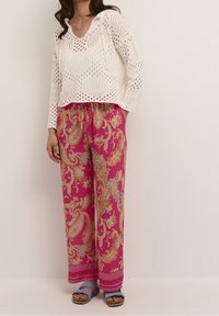 Biały dziergany sweter z dekoltem w serek i ażurowym wzorem, zestawiony z różowymi spodniami w orientalny wzór paisley oraz niebieskimi sandałami, z wiązaniem w talii.