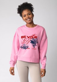 Lichtroze sweatshirt met lange mouwen, voorzien van bloemenafbeeldingen en de tekst "BE YOUR Self" in rood. Geribbelde, loszittende pasvorm.
