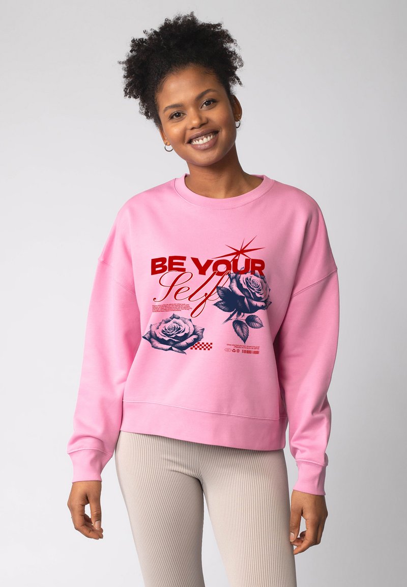 Lichtroze sweatshirt met lange mouwen, voorzien van bloemenafbeeldingen en de tekst "BE YOUR Self" in rood. Geribbelde, loszittende pasvorm.