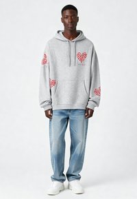 Sudadera gris con capucha y gráficos de corazones rojos en las mangas y el pecho, hecha de una mezcla de algodón, combinada con jeans de corte holgado en un tono azul claro.