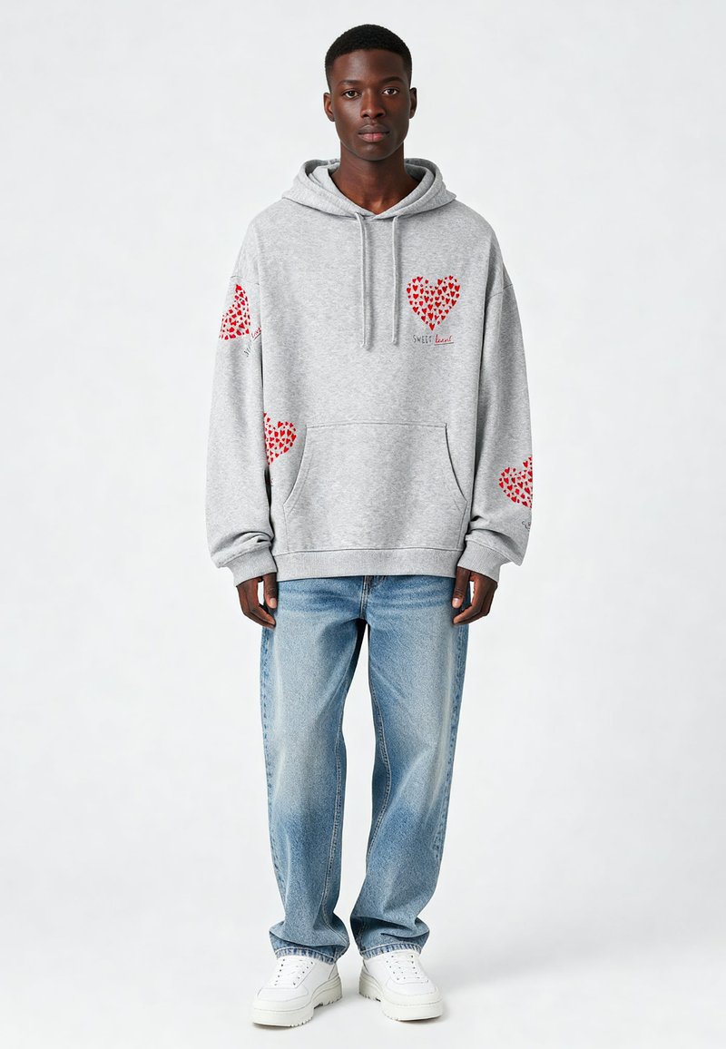 Sudadera gris con capucha y gráficos de corazones rojos en las mangas y el pecho, hecha de una mezcla de algodón, combinada con jeans de corte holgado en un tono azul claro.