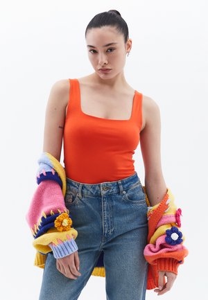 Rood-oranje aansluitend tanktop met hoge vierkante halslijn, gecombineerd met hoge taille, lichtblauwe denim jeans. Chunky, multicolore gebreide cardigan over de armen gedrapeerd.
