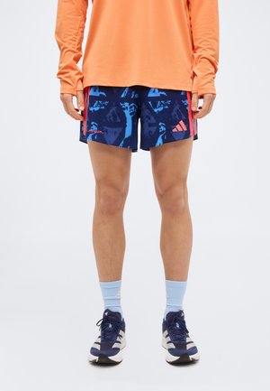 Persoană care poartă un tricou portocaliu cu mâneci lungi, pantaloni scurți atletici albaștri cu modele și dungi coral, șosete bleu deschis și încălțăminte sport bleumarin.