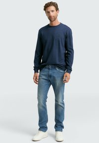 Marineblauwe longsleeve shirt met ronde hals, gecombineerd met lichtblauwe jeans en witte sneakers. Bevat een klein logo op de borst van het shirt.