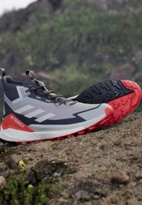 Szare i białe buty trekkingowe z czerwonymi akcentami oraz teksturowaną czarną podeszwą. Posiadają wysoki krój za kostkę oraz system szybkiego sznurowania.