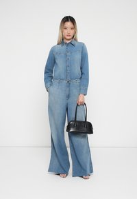 Denim jumpsuit with a collar, long sleeves, and button closure; light blue color; paired with a black leather handbag. Wide-leg fit.