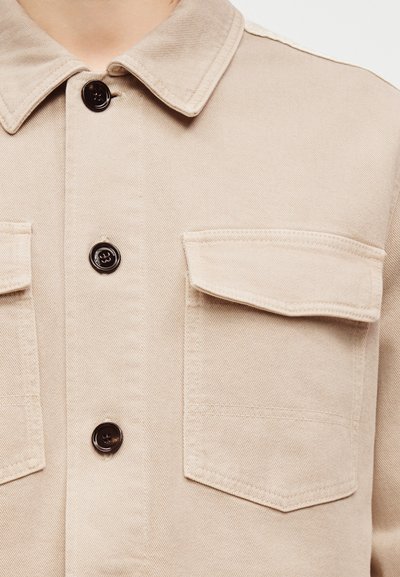 Μπεζ πουκάμισο με κουμπιά, με γιακά και δύο εμπρόσθιες τσέπες με καπάκι, εμφανίζοντας την περιοχή από το μέσο του στήθους έως τον λαιμό.