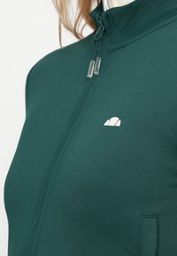 Tamnozeleni zip-up dukserica od glatkog, rastezljivog materijala. Ima visoki ovratnik i bijeli logo na lijevoj strani prsnog koša, s jasno vidljivim povlačnim patentnim zatvaračem.