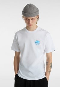 Vit bomull t-shirt med en ljusblå "VANS"-logotyp på bröstet. Modellen bär en grå stickad mössa och har synliga tatueringar på armen.