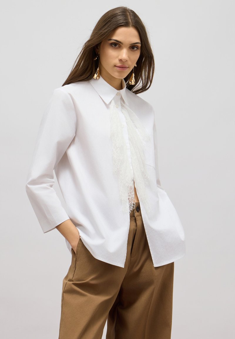 Camicia bianca a maniche lunghe, con colletto a punta, caratterizzata da un dettaglio in pizzo lungo la parte anteriore. Abbinata a pantaloni marroni a gamba larga.