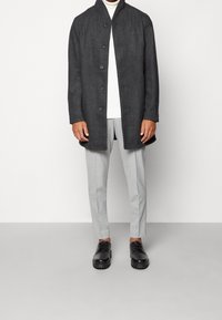 Szary wełniany płaszcz z wysokim kołnierzem, zapięciem na guziki i długimi rękawami, w połączeniu z jasnoszarymi spodniami i czarnymi sznurowanymi butami.