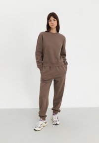 Ensemble de sport en tissu doux et marron composé d'un sweat-shirt à col ras du cou et d'un pantalon de jogging avec des poignets élastiques. Modèle portant des baskets épaisses.
