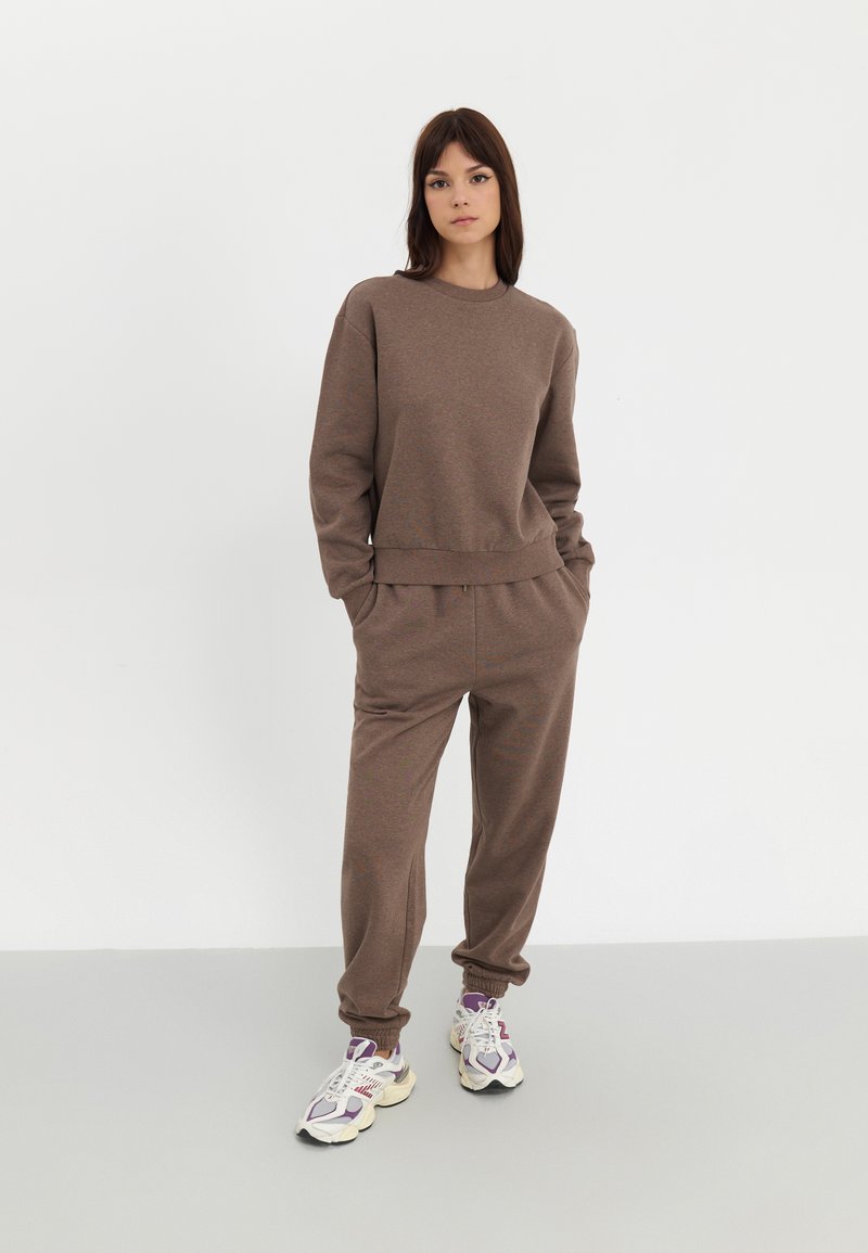 Ensemble de sport en tissu doux et marron composé d'un sweat-shirt à col ras du cou et d'un pantalon de jogging avec des poignets élastiques. Modèle portant des baskets épaisses.