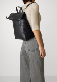 Mujer vestida con una blusa de canalé beige y jeans grises, llevando una mochila de cuero negro con un solapa de cremallera plegable.