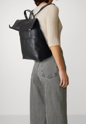 Donna con top a coste beige e jeans grigi, che porta uno zaino in pelle nera con un cappuccio a zip pieghevole.