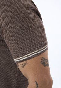 Chemise à manches courtes texturée marron avec un col côtelé. Le poignet présente un accent rayé blanc. Tatouage visible sur le bras.