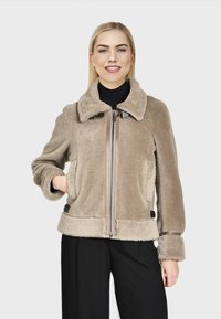 Jonge vrouw die glimlacht, gekleed in een beige fuzzy zip-up jack met grote kraag en zwarte broek, hand in de zak van het jack, tegen een effen achtergrond.
