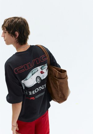 Jeune personne portant un t-shirt noir avec un graphique Honda Civic et un pantalon rouge, portant un grand sac en cuir marron sur une épaule.
