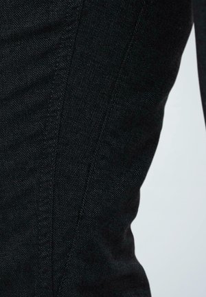 Schwarzer Denim-Stoff mit Fischgrätenmuster, der geschwungene Nähte und eine glatte Textur aufweist. Eine Nahaufnahme betont die Struktur des Stoffes.