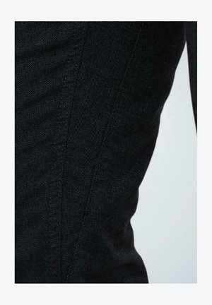 Zwarte denimstof met een visgraatpatroon, voorzien van gebogen naden en een gladde textuur. Close-up detail benadrukt de structuur van de stof.