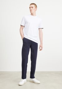 Weißes T-Shirt mit kurzen Ärmeln und schwarz-weißen gestreiften Bündchen, kombiniert mit dunkelblauen Hosen und weißen Sneakers, minimalistisches Design.