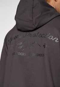 Siv hoodie s kapuco in velikim, embosiranim logotipom z gorami in besedilom na hrbtni strani. Mehak material z gladko teksturo.