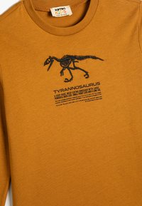 Musztardowa, długorękawowa koszulka z czarnym haftowanym szkieletem Tyrannosaurusa i opisującym tekstem poniżej. Wykonana z bawełny, gładka w dotyku.