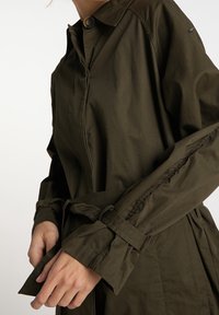 Lekka kurtka w oliwkowej zieleni z zapięciem na guziki, paskiem w talii i mankietami na rękawach. Posiada klasyczny kołnierz i minimalistyczne przeszycia.