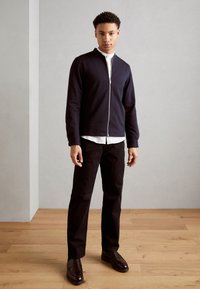 Mornarska zip-up sweatshirt s visokim ovratnikom, nošena čez belo srajco, v kombinaciji s črnimi hlačami in rjavimi usnjenimi čevlji na lesenem podu.