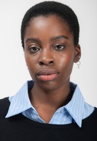 Zwarte trui over een lichtblauw, gestreept overhemd met kraag. Korte, gestructureerde haarstijl. Eenvoudige oorbellen. Neutrale achtergrond.