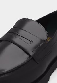 Μαύρο δερμάτινο μοκασίνι με λουράκι για κέρμα και λεπτομέρειες με οδοντωτή άκρη στη γλώσσα, εμφανιζόμενο από κοντινή γωνιακή όψη.