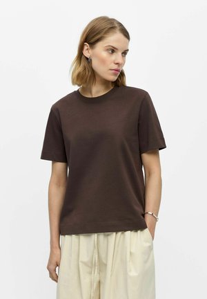 Femme aux cheveux courts châtain clair portant une chemise marron foncé à manches courtes et un pantalon beige ample sur un fond blanc uni.
