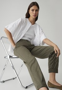 Vrouw met lang donker haar zittend op een zilveren klapstoel, gekleed in een wit T-shirt met korte mouwen, een wit hemdje, olijfgroene broek en bruine platte schoenen.