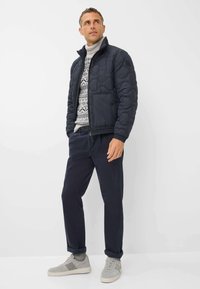 Marine gewatteerde jas met hoge kraag, grijs betegeld trui, donkere broek en lichtgrijze sneakers. Casual pasvorm met gestructureerde stof.