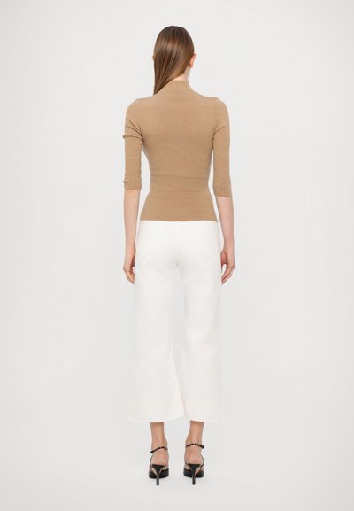 Haut marron ajusté à côtes avec col montant, associé à un pantalon large blanc. Modèle vu de dos, debout dans des sandales à talons noires.