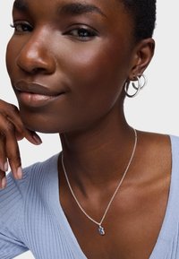 Srebrny naszyjnik z niebieskim wisiorkiem, noszony na jasnoniebieskiej, prążkowanej bluzce. Właścicielka ma na sobie kolczyki w kształcie kółek, a ręka spoczywa na szyi. Gładka faktura.