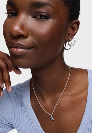 Collar de plata con un colgante azul, llevado sobre una blusa de rayas azul claro. Presenta pendientes de aro y una mano descansando sobre el cuello. Textura suave.