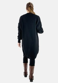 Svart oversized stickad klänning med ribbat mönster, nedhängda axlar och långa ärmar; kombinerad med svarta leggings och bruna knähöga stövlar.