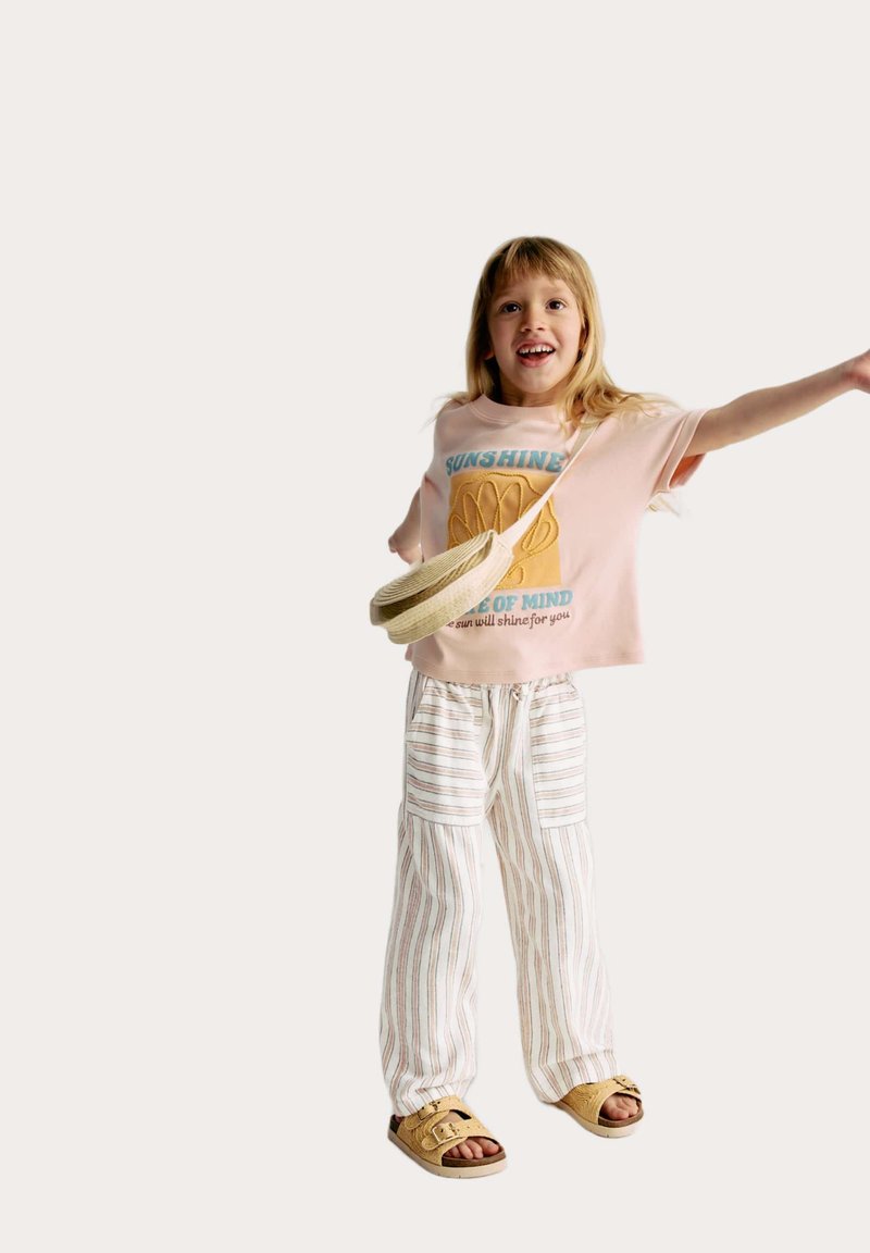 Ljusrosa T-shirt med blå och guld grafik, i kombination med vita randiga baggy-byxor. Bruna sandaler och en liten vävd väska kompletterar looken.