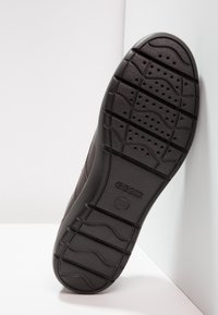 Zool van een donkerbruine Geox-schoen met een gestructureerde grip en perforaties, rechtop geplaatst tegen witte hoekwanden.
