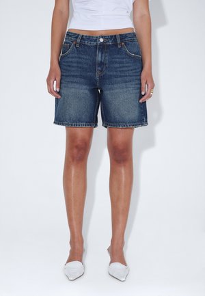 Kobieta nosząca ciemnoniebieskie jeansowe krótkie spodenki o średniej długości, biały krótki top i białe buty z ostrymi czubkami, stojąca na białym tle.