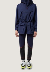 Marineblauwe lichtgewicht jas met een capuchon, elastische trekkoord taille en grote voorzak; gecombineerd met marineblauwe en zwarte slimfit broeken en felgekleurde sneakers.