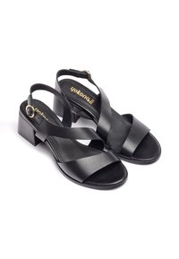 Sandalias de cuero negro con tacón ancho, con correas cruzadas y una correa ajustable en el tobillo con un hebilla dorada. Textura suave y diseño minimalista.