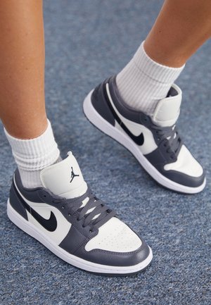 Osoba nosząca czarno-białe buty Nike Air Jordan i białe skarpetki do kostki na niebieskiej, teksturowanej podłodze.