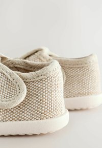 Gros plan de chaussures en tissu tissé beige avec bords arrondis et semelles en caoutchouc blanches sur un fond clair uni.