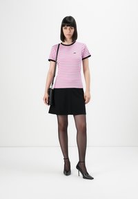 Frau steht in einem rosa-weißen gestreiften T-Shirt, schwarzem Rock, durchsichtigen schwarzen Strumpfhosen, schwarzen High Heels und einer schwarzen Umhängetasche.