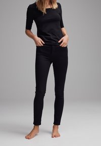 Woman wearing a black short-sleeve top and black skinny jeans standing barefoot with hands in pockets against a gray background.