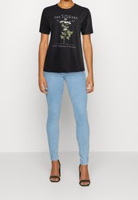 Camiseta de algodón negra con diseño gráfico floral y texto, combinada con jeans de mezclilla azul claro y sandalias de tacón negras.